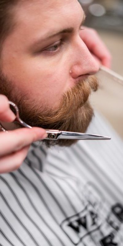 Close-up shot of a barber grooming a bearded man's beard and mustache with scissors.